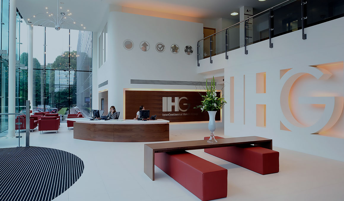 Two receptionists sit at the curved welcome desk in the lobby of the InterContinental Hotels Group corporate office. A large 3D version of the IHG logo serves as a dominant focal point.