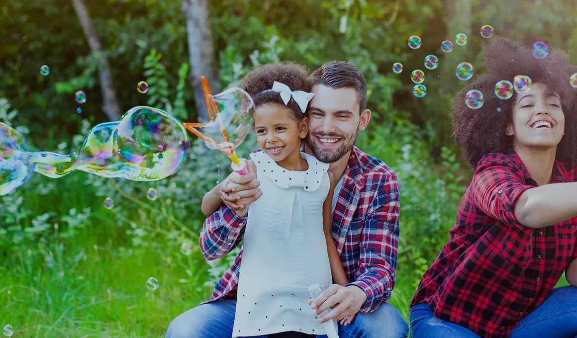 A husband and wife with their daughter, all smiling while using bubble wands outside.