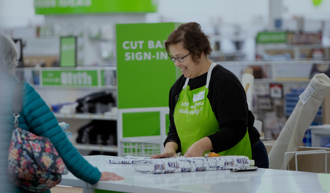 JOANN team member helping a customer at the cut bar, cutting fabric from a bolt, in the flagship Polaris store. She is wearing a bright green apron, smiling while focusing on her task.